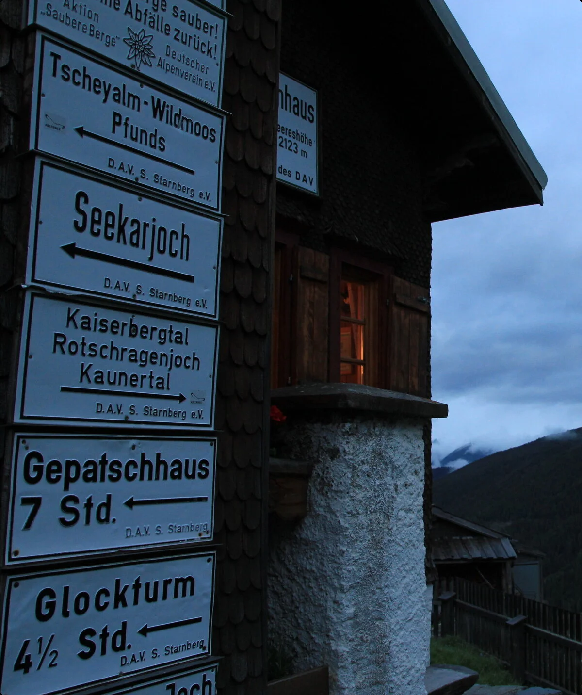 Wegbeschilderung an der Wand des Hohenzollernhauses bei düsterer Bewölkung mit Nebenhütte im Hintergrund | © DAV Starnberg