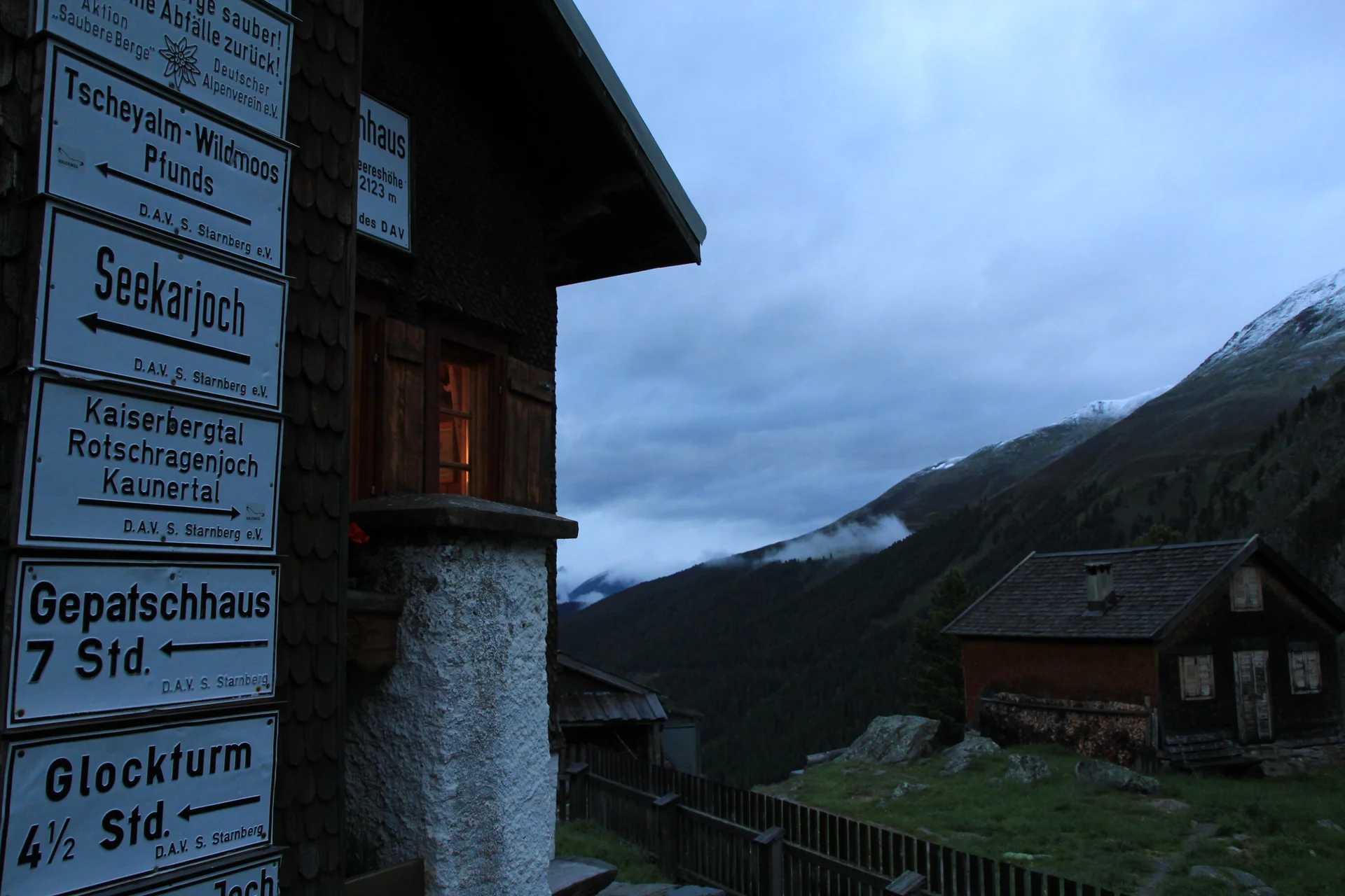 Wegbeschilderung an der Wand des Hohenzollernhauses bei düsterer Bewölkung mit Nebenhütte im Hintergrund | © DAV Starnberg